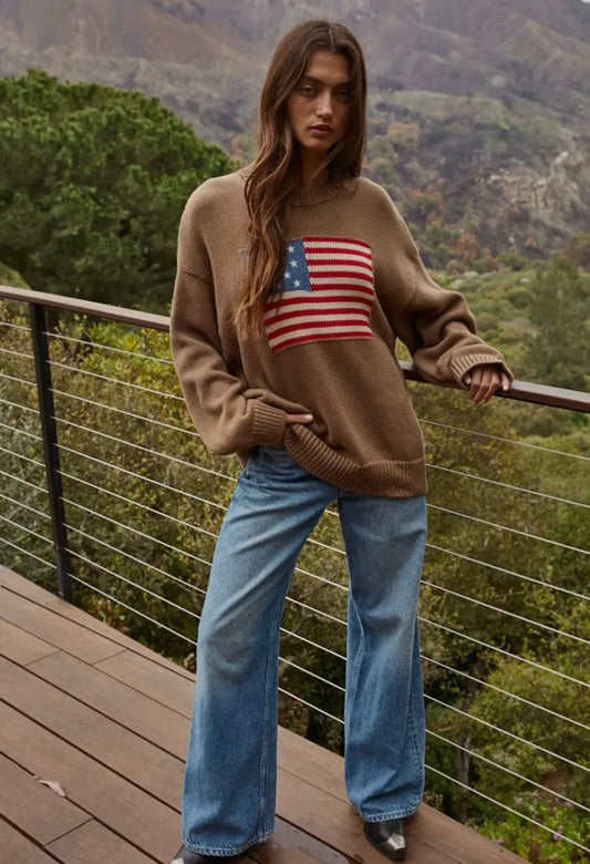 Person wearing a brown sweater with an American flag design, standing on a wooden deck with a scenic background.